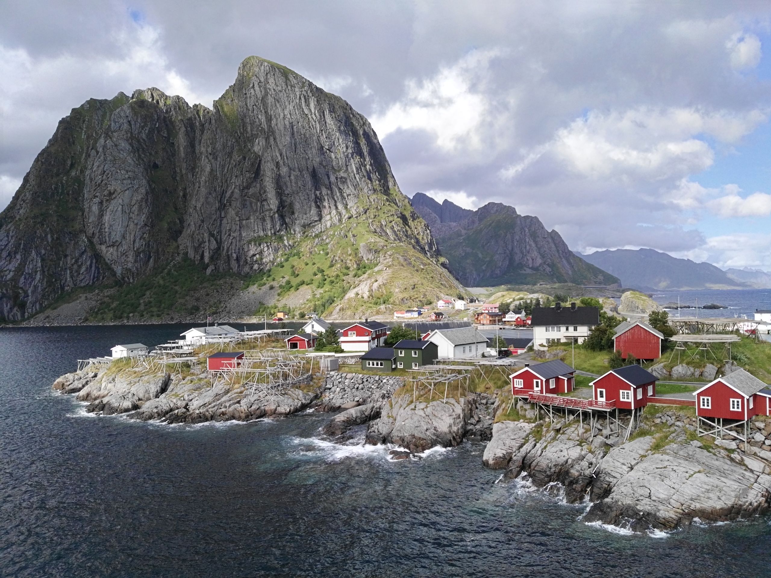 Piccolo villaggio costiero con case rosse e montagne imponenti sotto un cielo nuvoloso, Lofoten in bici, viaggi in bicicletta "Girolibero"